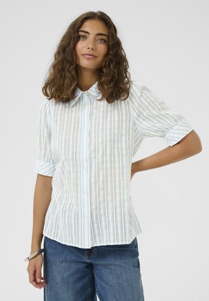 Mujer con cabello rizado y castaño, vestida con una camisa de rayas azul claro y blanca abotonada y jeans azules, de pie con una mano en la cadera.