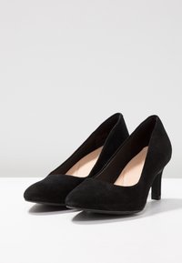 Talons hauts en suède noir avec un bout pointu et un talon fin. L'intérieur est doublé d'un matériau beige doux. Texture lisse sur toute la surface.