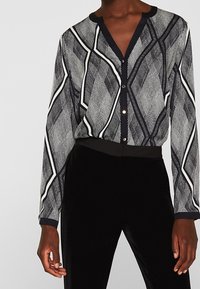 Donna che indossa una camicia a bottoni con un motivo bianco e nero, a maniche lunghe, infilata in pantaloni neri a vita alta.
