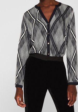 Vrouw in een zwart-wit gestreepte blouse met lange mouwen, ingestopt in zwarte high-waisted broeken.
