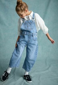 Lichtblauwe spijkeroveralls met verstelbare banden, wijde pijpen, voorzakken en zilverkleurige hardware, gecombineerd met een wit shirt en zwarte sneakers.