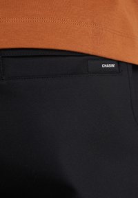 Svarta byxor med en slät yta, med en synlig logotagg märkt "CHASIN'" på midjan och en orangeklädd skjorta som delvis överlappar toppen.