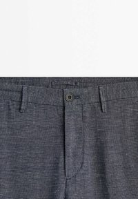 Gros plan sur un pantalon gris foncé texturé montrant une ceinture boutonnée avec passants de ceinture et braguette.