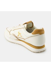 Sneaker bianco con tomaia in pelle liscia, accenti marroni e suola in gomma texturizzata. Presenta un logo sul lato e cuciture decorative.