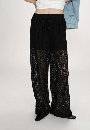 Pantalon large en dentelle noire avec des motifs floraux porté sur un short noir, associé à des chaussures noires pointues et un haut court blanc.