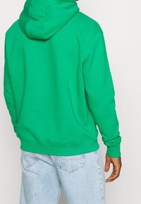 Sudadera con capucha verde de tejido de mezcla de algodón, con capucha ajustable y puños de canalé, combinada con unos vaqueros de mezcla de denim de color azul claro.
