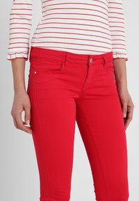 Jean en denim extensible rouge avec une coupe slim, doté d'une poche avant, d'une fermeture par bouton, et d'une texture lisse. Associé à un haut rayé.