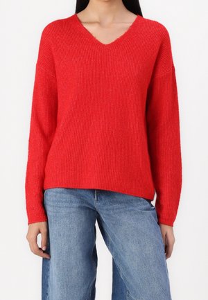 Frau trägt einen leuchtend roten, gerippten V-Ausschnitt-Pullover kombiniert mit locker sitzenden blauen Jeans, stehend vor weißem Hintergrund.
