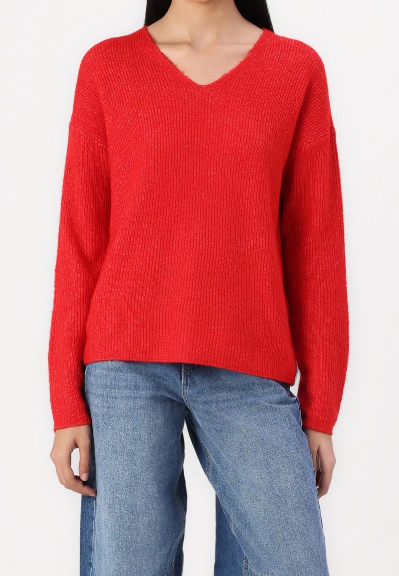 Femme portant un pull rouge vif à col en V côtelé associé à un jean bleu ample, debout sur un fond blanc.