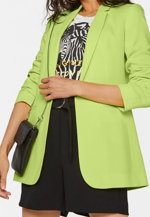 Femme portant un blazer vert lime, un short noir, un t-shirt blanc à imprimé zèbre et un sac bandoulière noir avec une lanière chaîne.