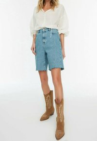 Mujer con blusa blanca holgada, bermudas de mezclilla azul claro de tiro alto y botas vaqueras de ante color canela con costuras blancas.