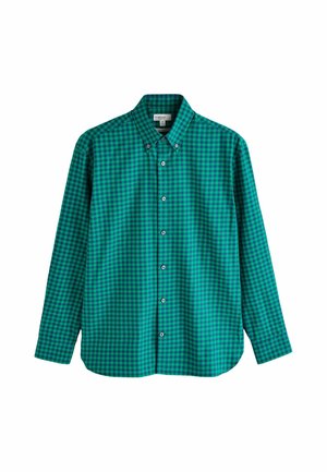 Jeune homme aux cheveux bouclés portant une chemise à carreaux verte et bleue à boutons, regardant vers sa gauche sur un fond uni.
