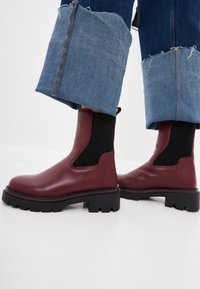 Stivaletti in pelle bordeaux con suola nera spessa, pannelli laterali elasticizzati e dettagli testurizzati; finitura liscia e punta arrotondata.