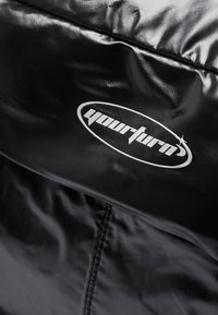 Crna podstavljena jakna s sjajnom teksturom i vidljivim šavovima. Sadrži istaknuti srebrni logo s natpisom "yourturn" na prednjoj strani.