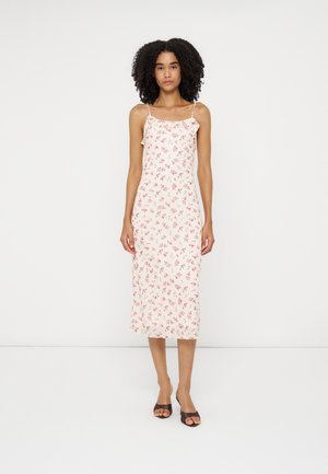 Kvinde med krøllet hår iført en hvid midi-kjole med pink blomsterprint og tynde stropper, stående mod en ensfarvet hvid baggrund.
