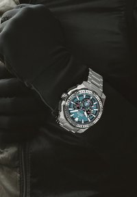 Reloj de acero inoxidable con un dial turquesa, acentos plateados, características de cronógrafo y una correa robusta, llevado en una muñeca con guantes.