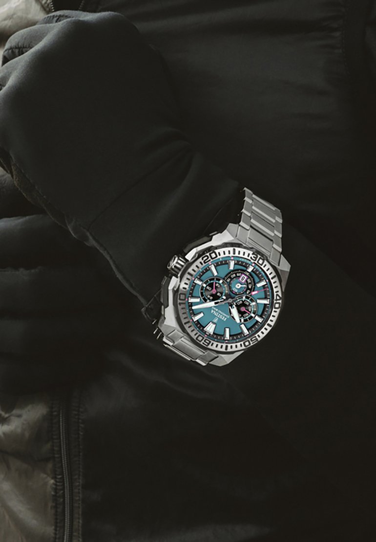 Reloj de acero inoxidable con un dial turquesa, acentos plateados, características de cronógrafo y una correa robusta, llevado en una muñeca con guantes.