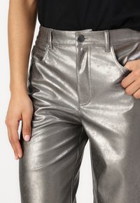 Metallic silver pants made from a shiny fabric, featuring front pockets, a button closure, and a smooth texture with slight wrinkles.