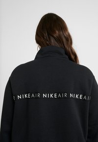 Svart sweatshirt med hög krage, prydd med ett stort vitt "NIKE AIR" logoband på baksidan. Mjukt, texturerat tyg.