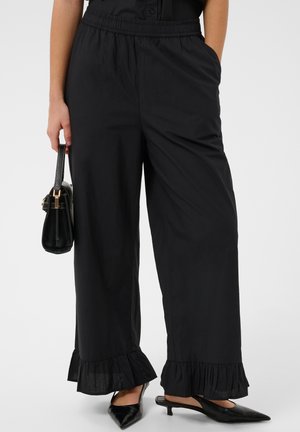 Kobieta nosząca czarne spodnie o szerokich nogawkach z marszczonymi brzegami, czarne spiczaste buty typu slingback, trzyma małą czarną torebkę przy boku.