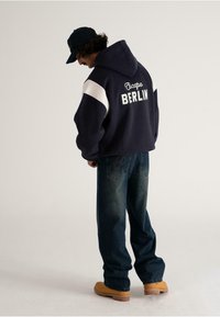 Person wearing navy hoodie with "Escape Berlin" text, dark jeans with embroidery, tan boots, and black cap, standing against a plain background.