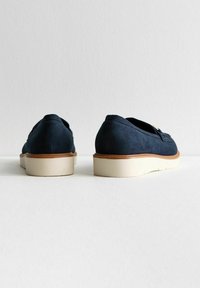 Chaussures slip-on en daim bleu marine avec une semelle en caoutchouc crème et des accents marron. Texture lisse avec des détails cousus sur les côtés.