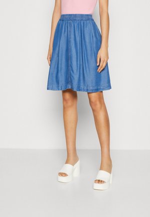 Kobieta ubrana w niebieską spódnicę dżinsową do kolan z elastycznym pasem, różową bluzkę oraz białe sandały na platformie z odkrytymi palcami, na jednolitym tle.