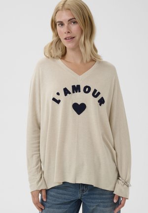 Blonde Frau trägt einen beigen V-Ausschnitt-Pullover mit "L'AMOUR" und einem schwarzen Herz vorne sowie blaue Jeans.