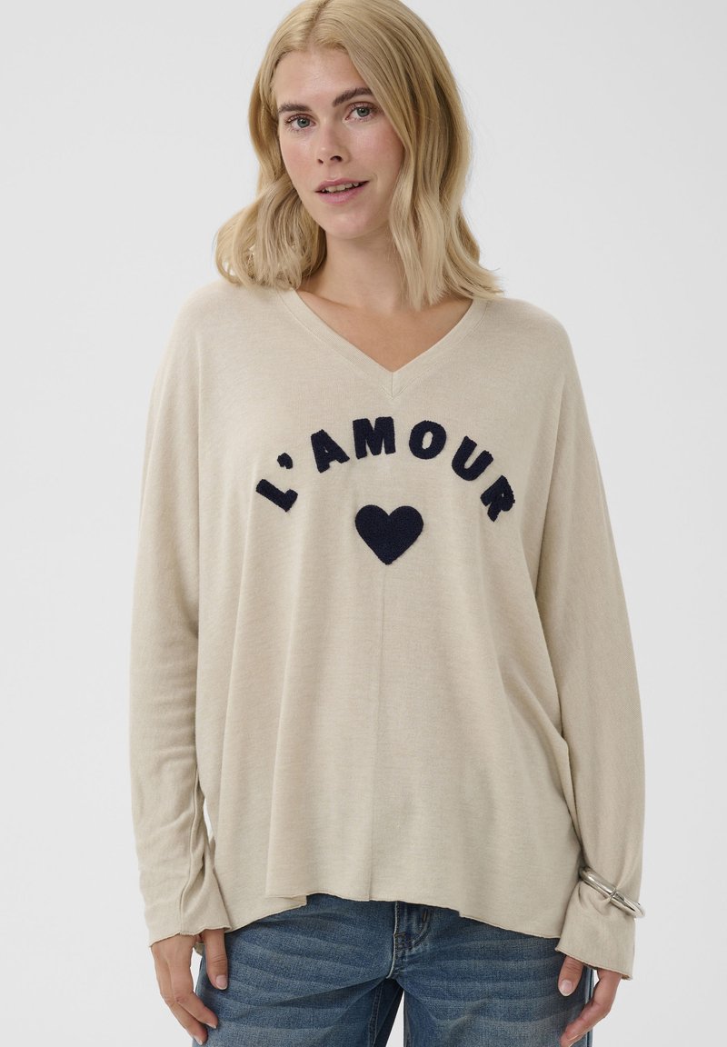 Femme blonde portant un pull beige à col en V avec "L'AMOUR" et un cœur noir imprimé sur le devant, ainsi qu'un jean bleu.