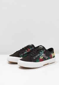 Mustat matalat tennarit, joissa on valkoiset pohjat, kukkakuvio ja "Karl" kirjailtuna sivuille, esitettynä yksinkertaista valkoista taustaa vasten.