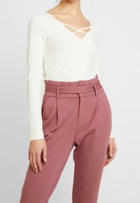 Valkoinen pitkähiha bodysuit syvällä V-pääntiellä ja ristikkäissuunnassa olevilla olkaimilla, yhdistettynä rakenteellisiin, korkeavyötäröisiin, ruusupinkkeihin housuihin.