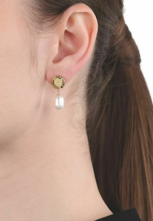 Boucle d'oreille en or avec cercle gravé et perle blanche pendante sur l'oreille d'une femme aux cheveux bruns tirés en arrière.