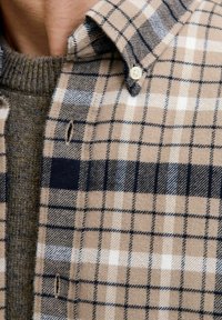 Beżowa i granatowa koszula w kratę z guzikami nałożona na szary sweter dzianinowy, ukazująca kołnierz i klatkę piersiową osoby.