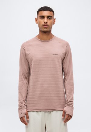 Homme portant un haut côtelé à manches longues couleur mauve clair avec des ouvertures pour les pouces, debout devant un fond blanc uni.