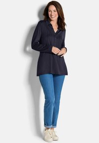 Marineblaues Hemd mit langen Ärmeln, Details auf der Vorderseite, kombiniert mit hellblauen Jeans und weißen Sneakers. Glatter Stoff, lässige Passform.