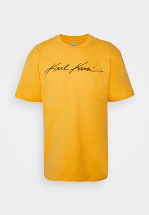 Gelbes T-Shirt mit kurzen Ärmeln und schwarzem kursivem Schriftzug "Karl Kani" über der Brust vor einem einfarbig hellen Hintergrund.