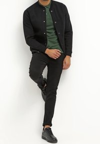 Man som bär en svart jacka, grön pikétröja, svarta byxor och svarta sneakers, poserar med ett ben lyft mot en vit vägg.