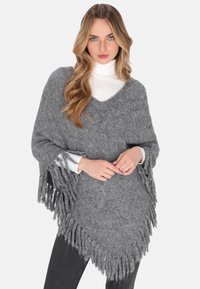 Frau in einem grauen, gestrickten Poncho mit Fransen über einem weißen Rollkragenpullover und schwarzen Hosen, stehend vor einem schlichten Hintergrund.