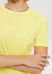 T-shirt giallo a maniche corte in cotone morbido, colletto a girocollo con maniche arrotolate. Accessoriato con una collana in argento e un anello semplice.