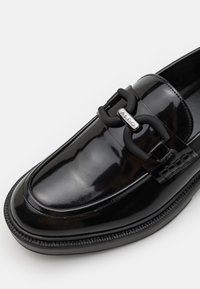 Mocassin en cuir noir avec une finition brillante, comportant une chaîne décorative proéminente et un accent Sigle ALDO sur la sangle.