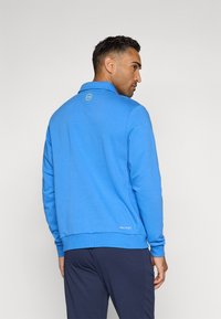 Sweatshirt bleu avec col, texture douce et poignets côtelés. Présente un logo dans le dos et un ourlet droit. Porté avec un pantalon bleu marine.