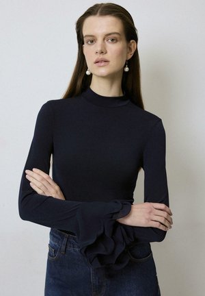 Navy long-sleeve top with a high neck and ruffle accents at the cuffs, paired with dark denim jeans, smooth fabric, and fitted design.