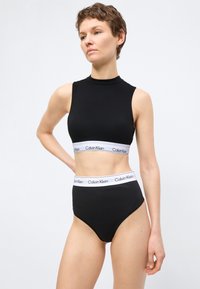 Γυναίκα που φοράει μαύρο αμάνικο κοντό τοπ και σλιπ ψηλόμεσο με λευκές ελαστικές ταινίες Calvin Klein, στέκεται μπροστά σε απλό λευκό φόντο.