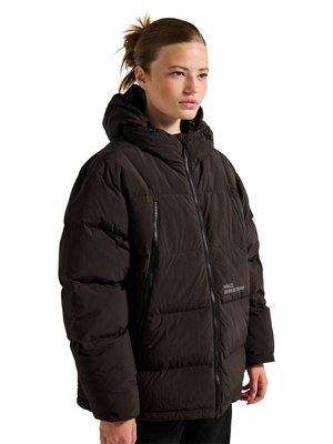 Ung kvinde med samlet hår iført en sort polstret hættejakke med forreste lynlås og sidelommer, stående og kigger fremad.