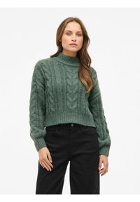 Pull en maille vert avec un col haut, présentant des motifs en torsades et des manches larges, associé à un pantalon noir.