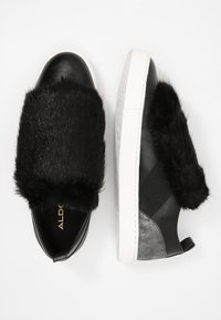 Černé slip-on tenisky s tlustým černým umělým kožíškem přes vršek, bílou gumovou podrážkou a stříbrnými tečkami na zadním podpatku.