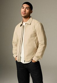 Veste légère beige avec fermeture éclair, col, et poches latérales, associée à un t-shirt blanc et un jean foncé. Texture lisse.
