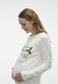 Hvid mellemlangærmet graviditetstrøje med farverig tekst og kristtorn-design. Blødt stof, fitted snit, fremhæver den runde maveform.