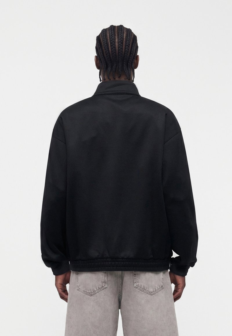 Person mit geflochtenem Haar, von hinten gesehen, trägt eine schlichte schwarze Jacke und hellgraue Hose vor weißem Hintergrund.