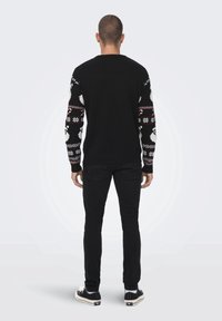 Maglione nero con maniche a fantasia festiva in bianco, rosso e grigio. Indossato con jeans neri e scarpe nere, visto da dietro.
